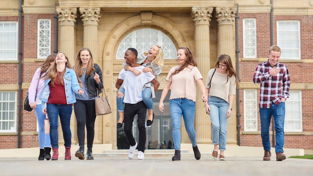 A large groups of students walk and laugh together outside of the main building