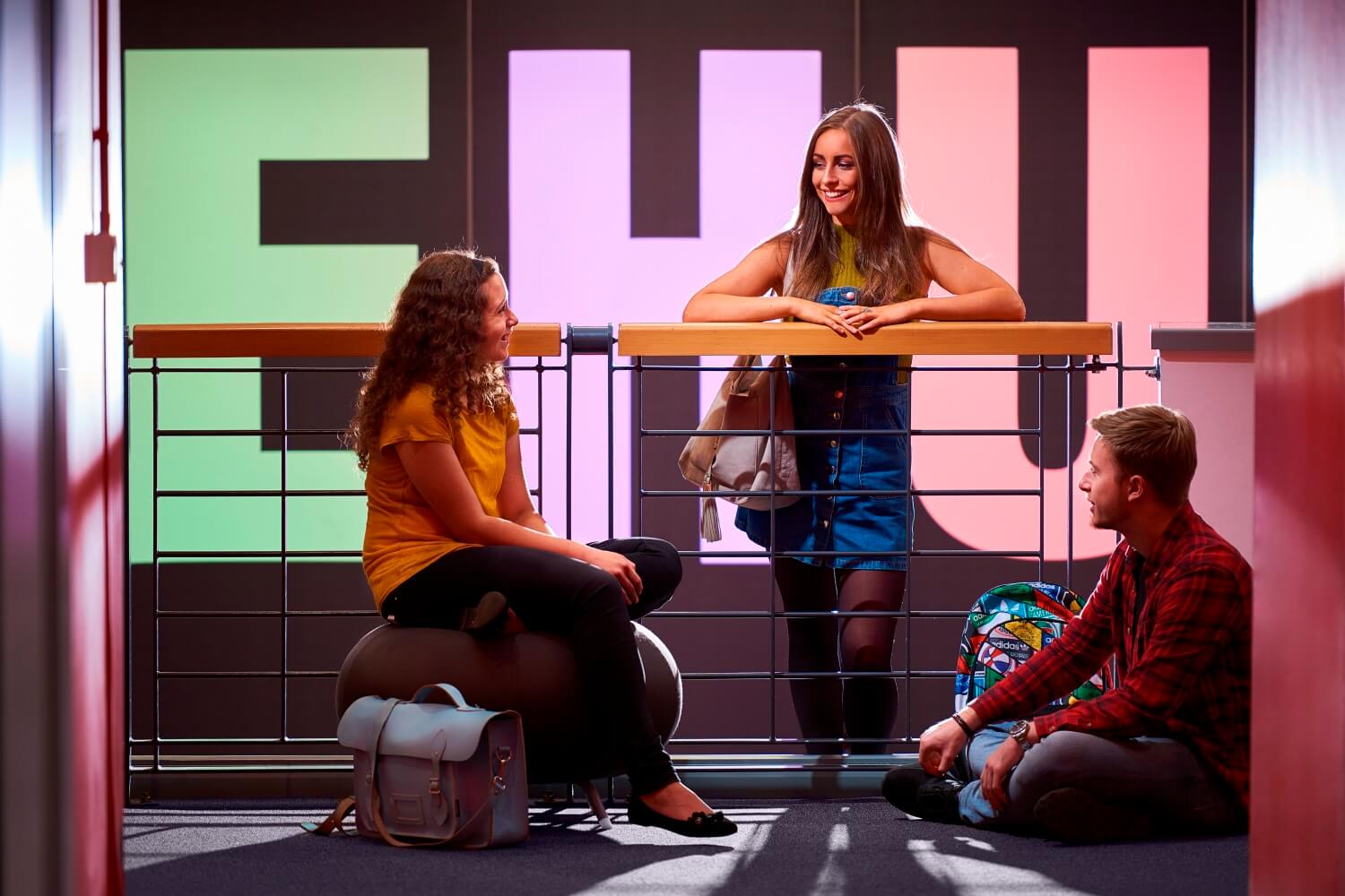 Three students sat in the foyer of the Arts Centre with EHU illuminated in large letters on the wall behind them.