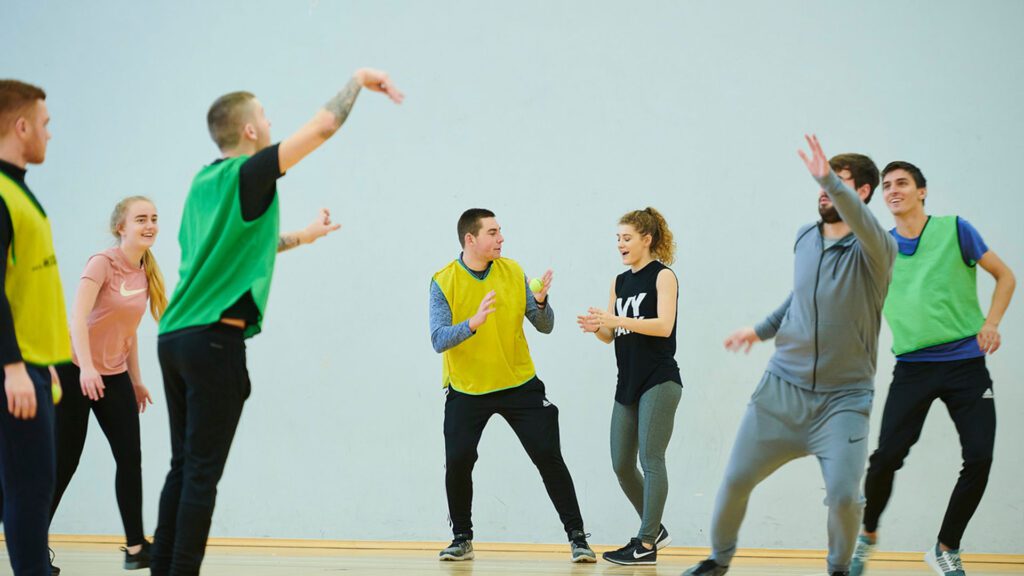 A group of sports students throwing a tennis ball in the gym. There are two teams of students with half of the students wearing yellow or green sports bibs.