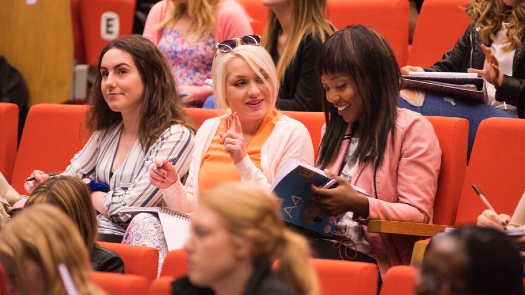 A group of nursing students laugh and smile sat in a row at a lecture theatre