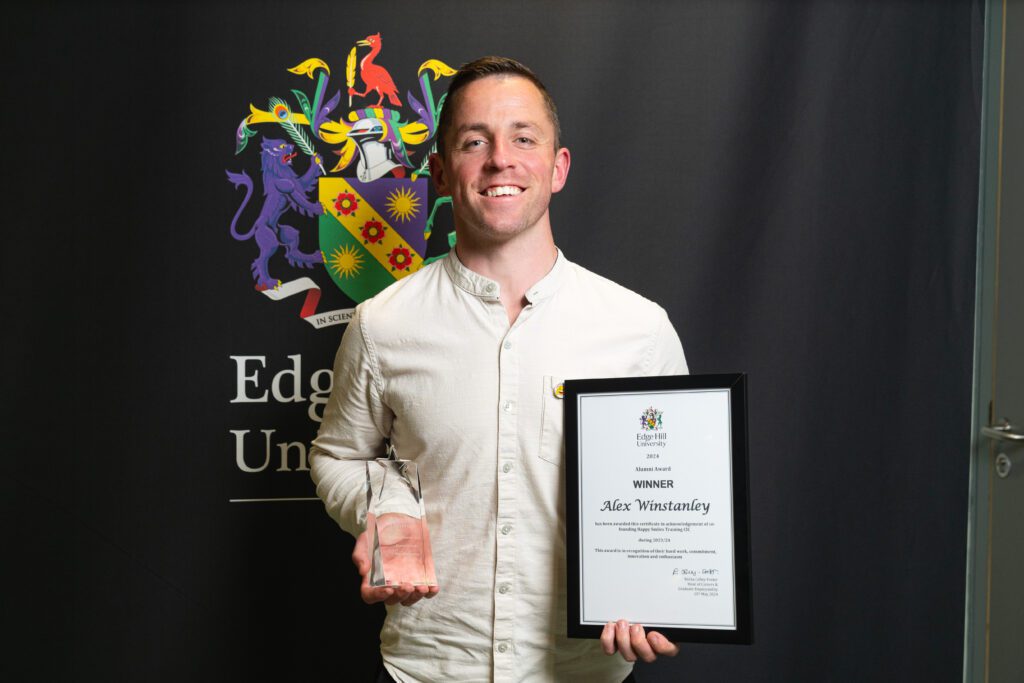 Alex Winstanley with their glass award and framed certificate in front of the Edge Hill University crest.