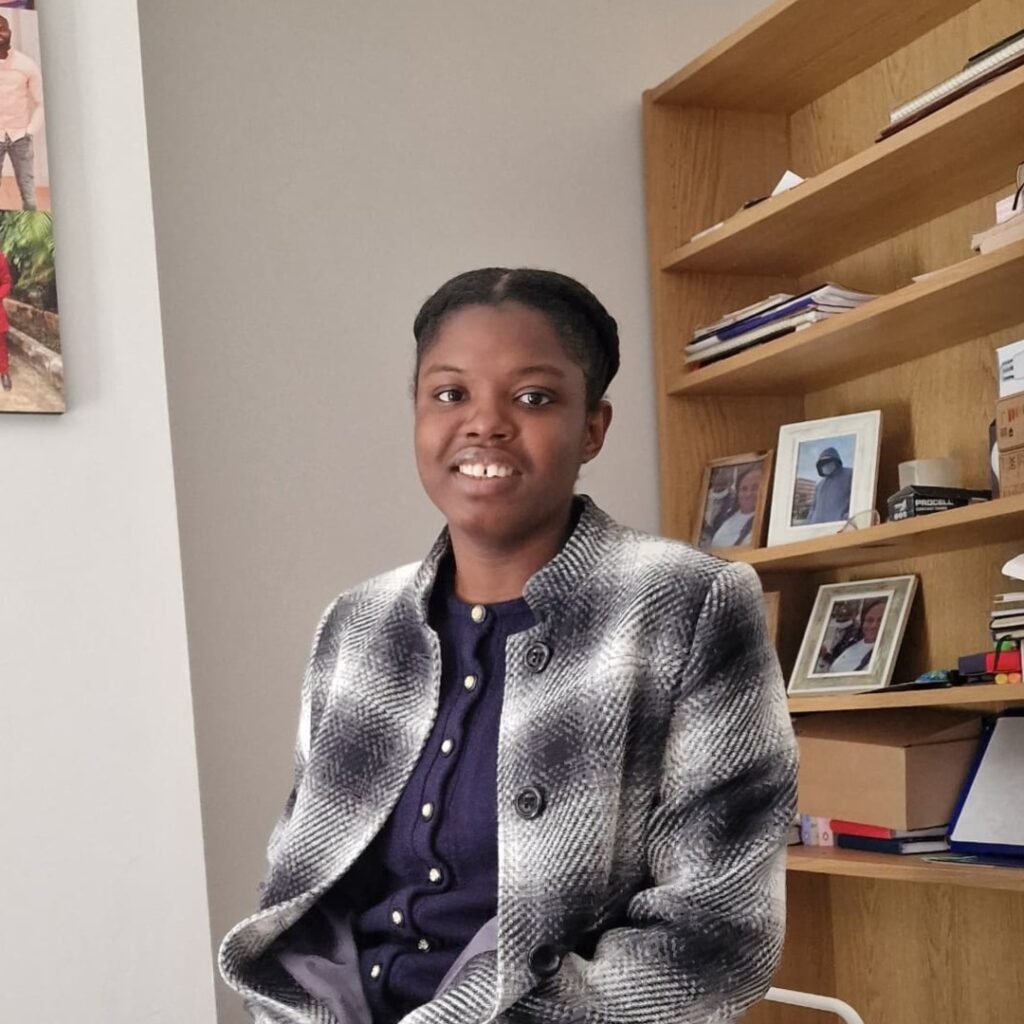 Racheal smiles while sitting on a chair arm at home with a busy bookcase behind her.