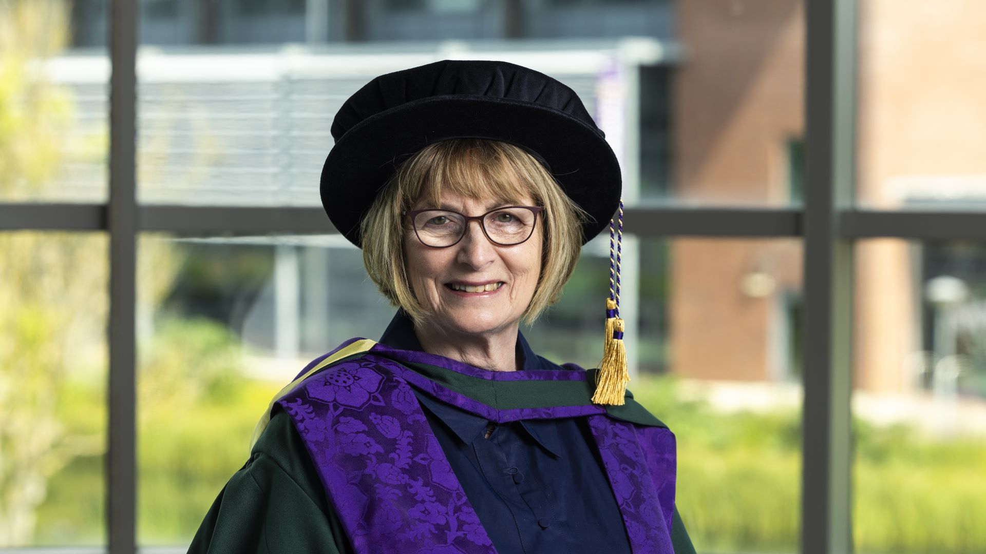 Dame Louise Ellman wears a black hat with a gold tassel and purple robes.