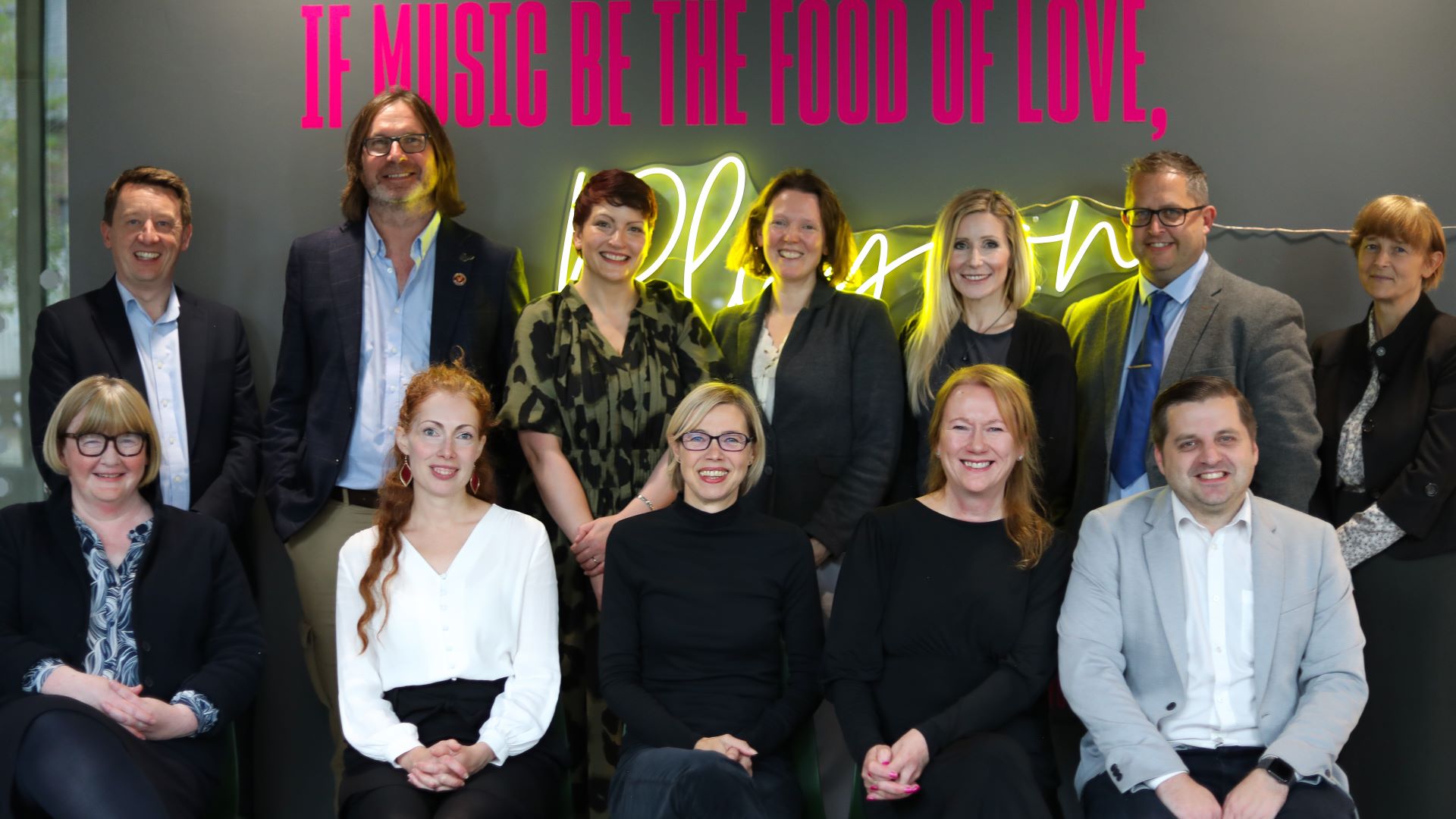 12 people stand and sit in front of a neon sign saying 