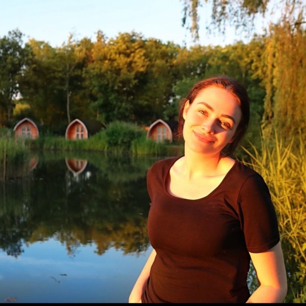 Jade Ainsworth stands in late afternoon sunshine with a lake and wooden huts in the background.