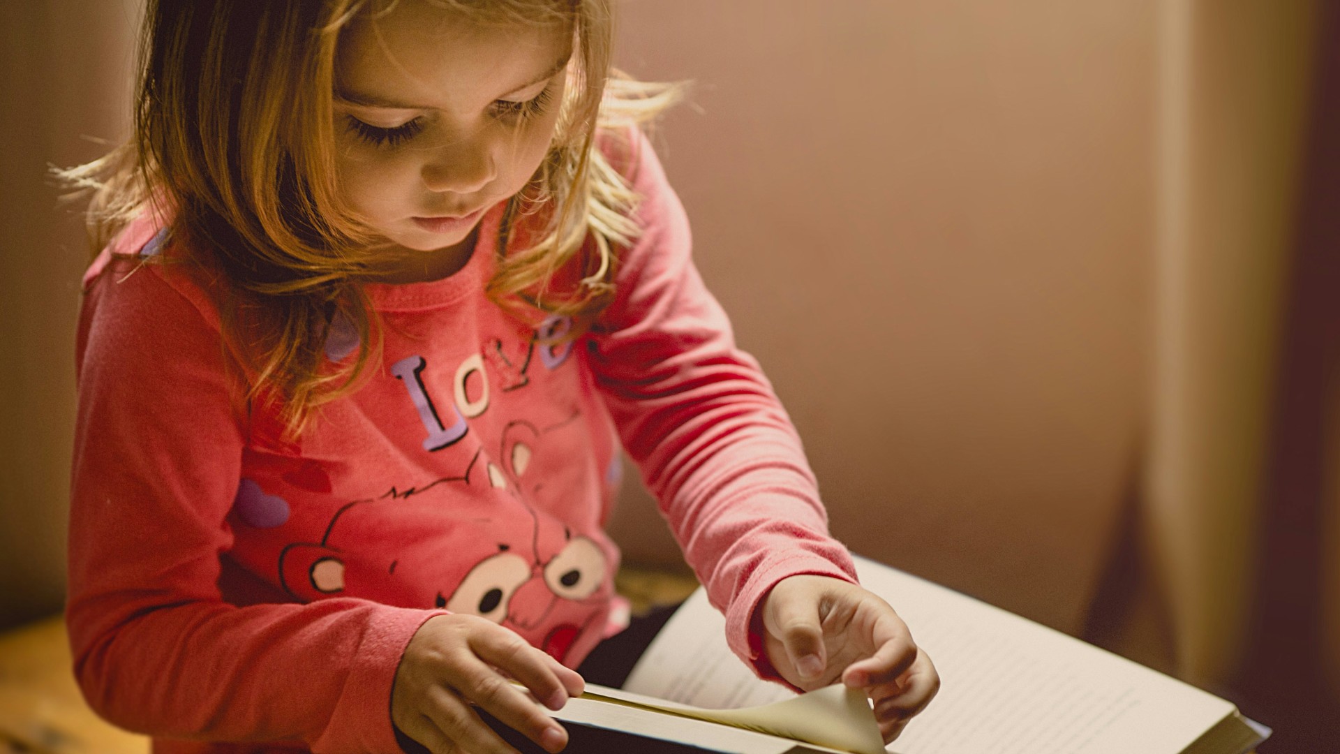 A young girl sits quietly, engrossed in reading a book, with a look of concentration on her face.