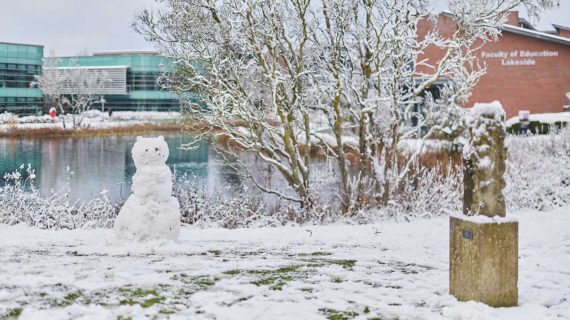 A photo of the Edge Hill western campus lake covered in snow and ice with a friendly bear-shaped snowman in the foreground.