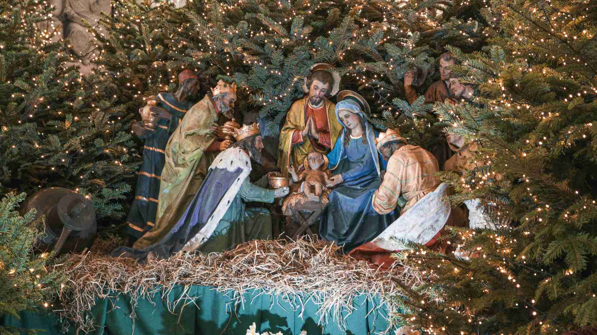 A traditional nativity scene with statues of Mary, Joseph, baby Jesus, and the three wise men, surrounded by shepherds and animals. The backdrop features twinkling lights and trees.