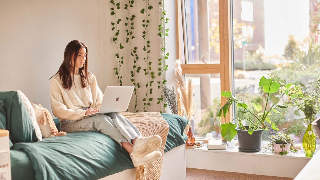 A student sitting on her bed using her laptop in her accommodation room.