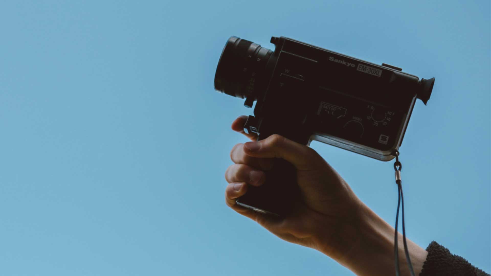 A hand holds a vintage Sankyo EM-30XL video camera in front of a sky blue blackground